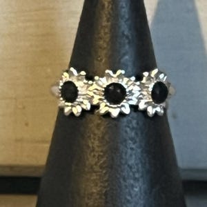 Peut inclure: Bague en argent ornée de trois motifs de tournesols avec des centres noirs. La bague est présentée sur un présentoir noir en forme de cône, mettant en valeur ses détails floraux complexes et sa finition polie. Un bijou unique et accrocheur.