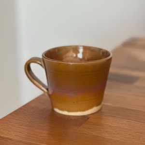 Op de afbeelding: Een keramische mok met een glanzende afwerking, voorzien van een warm okerbruin glazuur dat overgaat in een subtiele roodpaarse tint nabij het midden, met een eenvoudige, ongeglazuurde lichtbeige basis en een gebogen handvat aan de linkerzijde.