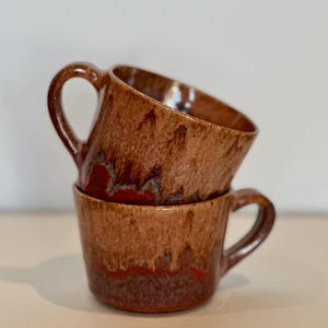 Puede incluir: Dos tazas de cerámica apiladas con un esmalte marrón y rojo. Las tazas tienen forma redondeada y un asa. El esmalte tiene un efecto de goteo, creando una textura y variación de color únicas.