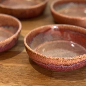 Könnte beinhalten: Vier handgefertigte Keramikschalen mit rustikalem, glasiertem Finish. Die Schalen haben eine rotbraune Außenseite, die in einen dunkleren roten und braunen Innenraum übergeht. Die Schalen sind auf einer Holzoberfläche angeordnet.