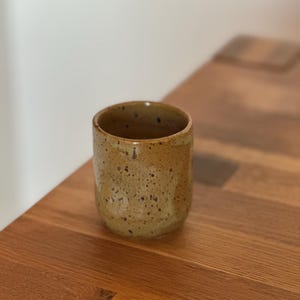 Può includere: Una tazza cilindrica in ceramica con una smaltatura maculata beige e marrone. La superficie esterna presenta un aspetto strutturato e terroso con macchie marrone scuro distribuite sul corpo, mentre l'interno rimane liscio e aperto.