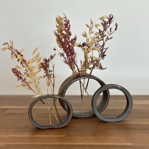 Op de afbeelding: Drie decoratieve metalen ringvazen, elk met gedroogde bloemen en grassen. De vazen zijn in een rij gerangschikt, met de grootste in het midden en de kleinste rechts. De gedroogde planten zijn in tinten van bruin, beige en bordeaux.