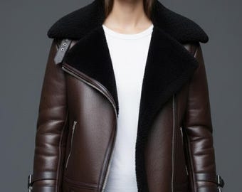Puede incluir: Chaqueta de cuero marrón con cuello y forro de borrego negro. La chaqueta tiene cierre de cremallera, múltiples bolsillos y correas ajustables. La modelo lleva una camiseta blanca y pantalones negros.