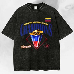 May include: Black acid wash t-shirt featuring a baseball trophy design with the text "2026 World Baseball Classic Champions" and the Venezuelan flag. The word "Venezuela" is also displayed.
