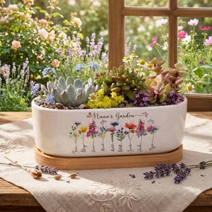 Peut inclure: Un pot de fleurs en céramique blanche avec l'inscription "Nana's Garden" et des illustrations florales, rempli de diverses succulentes et petites fleurs. Le pot repose sur un plateau en bambou, avec des brins de lavande à proximité.