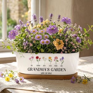May include: A white ceramic planter filled with a variety of colorful flowers, including purple lavender, yellow daisies, and orange blooms. The planter has the text "GRANDMA'S GARDEN" and names below illustrated flowers. It sits on a wooden tray.