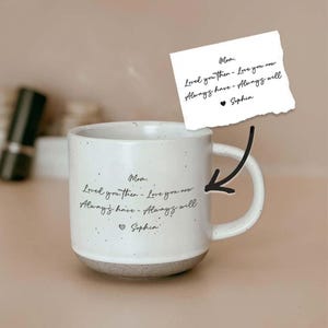 Könnte beinhalten: Weißer Keramikbecher mit gesprenkeltem Design und grauem Boden. Der Becher hat einen gebogenen Griff und handschriftlichen Text mit der Aufschrift "Mama, Habe dich damals geliebt - Liebe dich jetzt, Immer da - Immer da, Sophia."