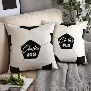 Op de afbeelding: Twee decoratieve sierkussens met een voetbalontwerp. De kussens zijn wit met zwarte vijfhoeken, die een voetbal nabootsen. Elk kussen heeft de naam "Charles" en het nummer "09" gedrukt op een zwarte vijfhoek.