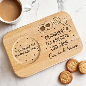 May include: A wooden tea and biscuit serving board with engraved text and cookie designs. The text reads "Grandma's Tea & Biscuits Love From Olivia & Henry" and "If you can read this Grandma needs a refill!!". A white mug of tea and biscuits are also present.