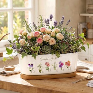 May include: A white ceramic planter filled with roses, lavender, and succulents. The planter has the text "Grandma's Garden" and floral illustrations. It sits on a wooden base, perfect for a kitchen or windowsill.