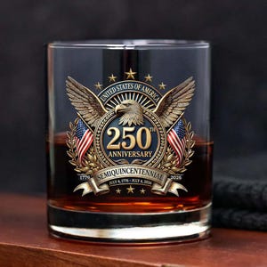 May include: A rocks glass with a patriotic design commemorating the 250th anniversary of the United States. The glass features an eagle, American flags, and the text "250th Anniversary" and "Semiquincentennial". The glass is filled with a dark amber liquid.