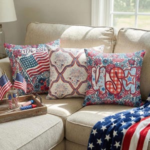 May include: Four decorative throw pillows with patriotic designs rest on a beige sofa. The pillows feature red, white, and blue patterns, including flags, fireworks, and floral motifs. A matching blanket with stars and stripes is draped on the sofa.