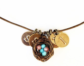 May include: A brown leather necklace with multiple charms, including a small bird's nest with blue and pink beads, and gold-toned metal charms with the letters 'S' and 'I'.