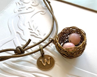 May include: A brown leather cord necklace with a brass charm in the shape of a bird's nest. The nest contains two pink glass beads. The charm is attached to a brass pendant with the letter 'W' on it.