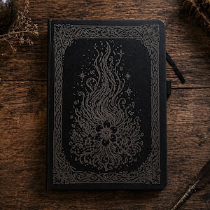 Peut inclure: Journal noir avec un motif floral et de flammes en relief. La couverture présente une bordure détaillée et un signet en ruban. Le journal est posé sur une surface en bois avec une plume.