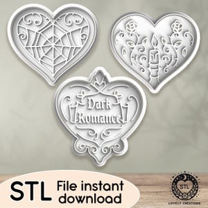 Puede incluir: Tres cortadores de galletas en forma de corazón blancos con diseños intrincados. El primer cortador presenta un diseño de telaraña, el segundo tiene un diseño de vela y flores, y el tercero tiene el texto "Dark Romance" con un borde decorativo.