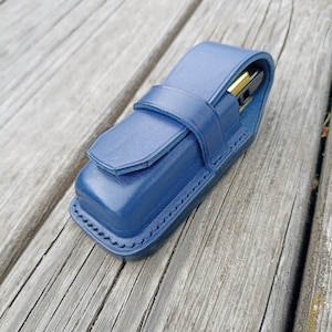 Op de afbeelding: Een blauwe leren etui met een klepsluiting en een riem. De rechthoekige etui heeft afgeronde randen en zichtbare stiksels. Een goudkleurige pen is gedeeltelijk zichtbaar in de etui.