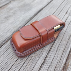 Op de afbeelding: Een bruine leren etui met een riemsluiting. De rechthoekige etui heeft een gestikte rand en een flap aan de bovenkant. Een goud-zwart object is gedeeltelijk zichtbaar aan de binnenkant.