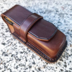May include: A brown leather case with a flap and strap closure. The rectangular case has rounded edges and a stitched border. The leather has a rich, textured appearance, and the strap secures the top flap. The case appears to be designed to hold a small object.