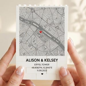 Puede incluir: Un bloque blanco con un mapa de París y un corazón rojo que marca una ubicación. El texto "ALISON & KELSEY" está debajo del mapa, junto con "EIFFEL TOWER" y coordenadas. También hay una fecha impresa en el bloque.