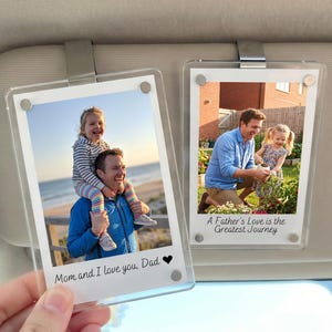 Könnte beinhalten: Zwei transparente Acryl-Fotorahmen, die an einer Sonnenblende befestigt sind. Jeder Rahmen enthält ein Farbfoto. Eines zeigt einen Mann mit einem Kind auf den Schultern, das andere einen Mann und ein Kind beim Gärtnern. Auf den Rahmen steht: "Mama und ich lieben dich, Papa" und "Die Liebe eines Vaters ist die größte Reise."