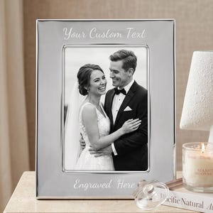 Puede incluir: Un marco de fotos rectangular de metal plateado con acabado pulido. El borde superior está grabado con el texto "Your Custom Text" y el borde inferior con "Engraved Here". Se muestra una fotografía de boda en blanco y negro.