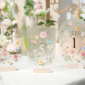 May include: Three clear acrylic table number signs with floral designs. Each sign has a different shape and features a number: 7, 5, and 1. The signs are decorated with watercolor flowers in pink, yellow, and blue, and sit on small wooden bases.