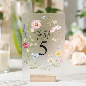 May include: A frosted acrylic table number with a floral design. The number 5 is in a bold black font, with the word "Table" in script above it. The flowers include daisies, lavender, and other colorful blooms. The sign is held by a small wooden base.