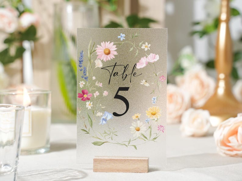 May include: A frosted acrylic table number with a floral design. The number 5 is in a bold black font, with the word "Table" in script above it. The flowers include daisies, lavender, and other colorful blooms. The sign is held by a small wooden base.