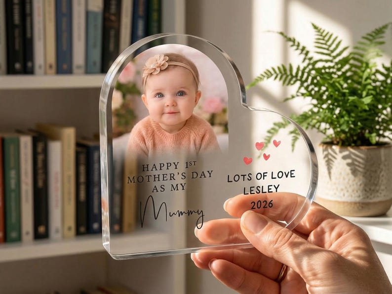 Puede incluir: Un marco de fotos acr&iacute;lico en forma de coraz&oacute;n con la foto de un beb&eacute;. El marco tiene el texto "Happy 1st Mother's Day as my Mummy" y "Lots of Love Lesley 2026" con peque&ntilde;os corazones rojos. Una mano sostiene el marco.