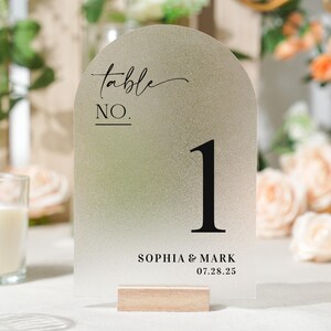 May include: A frosted acrylic table number with an arched top. The number one is in bold black font. The words "table NO." are in a script font. The names "SOPHIA & MARK" and the date "07.28.25" are printed at the bottom. The sign is held by a wooden base.