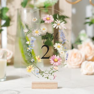 May include: Clear acrylic table number with a floral design. The number 2 is displayed in a large, bold font. The floral design includes pink, white, yellow, and blue flowers, with green leaves. The base is made of light-colored wood.