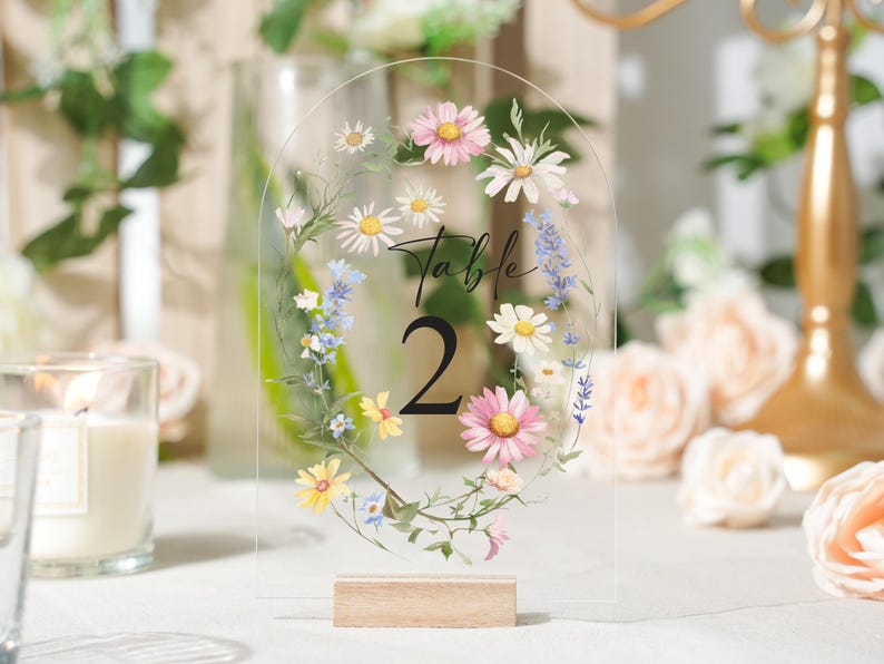 May include: Clear acrylic table number with a floral design. The number 2 is displayed in a large, bold font. The floral design includes pink, white, yellow, and blue flowers, with green leaves. The base is made of light-colored wood.