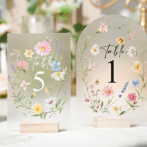 May include: Two frosted acrylic table number signs with floral designs. One sign is rectangular with the number 5, the other is arched with the number 1. Both signs have wooden bases and are decorated with pink, white, and blue flowers.