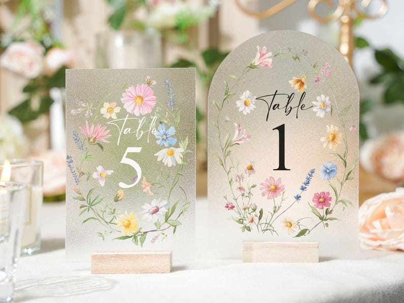 May include: Two frosted acrylic table number signs with floral designs. One sign is rectangular with the number 5, the other is arched with the number 1. Both signs have wooden bases and are decorated with pink, white, and blue flowers.