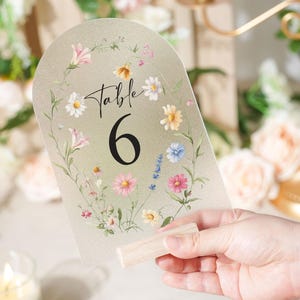May include: A clear acrylic table number with a floral design. The number 6 is in a bold black font. The floral design includes pink, yellow, and blue flowers. The table number is held by a wooden base.