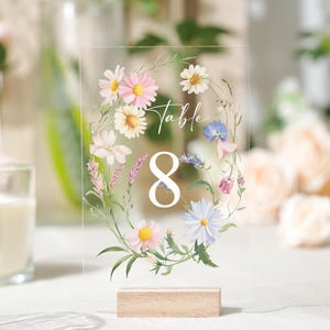 May include: Clear acrylic table number 8, adorned with a floral wreath of daisies, pink, blue, and purple flowers. The number is displayed on a wooden base, perfect for weddings or events.