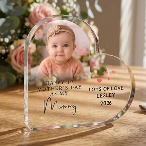 Puede incluir: Un marco de fotos acr&iacute;lico transparente en forma de coraz&oacute;n con la foto de un beb&eacute;. El marco tiene el texto "HAPPY 1ST MOTHER'S DAY AS MY Mummy" y "LOTS OF LOVE LESLEY 2026" con acentos de coraz&oacute;n rosa. Un ramo de flores est&aacute; en el fondo.