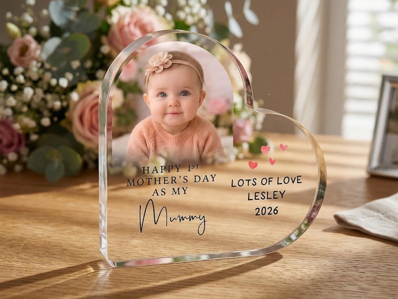 Puede incluir: Un marco de fotos acr&iacute;lico transparente en forma de coraz&oacute;n con la foto de un beb&eacute;. El marco tiene el texto "HAPPY 1ST MOTHER'S DAY AS MY Mummy" y "LOTS OF LOVE LESLEY 2026" con acentos de coraz&oacute;n rosa. Un ramo de flores est&aacute; en el fondo.