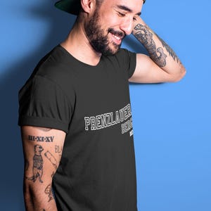 Könnte beinhalten: Schwarzes T-Shirt mit dem weißen Text "PRENZLAUER BERG", getragen mit einer schwarzen Baseballkappe. Das T-Shirt hat hochgekrempelte Ärmel. Der Hintergrund ist einfarbig blau.