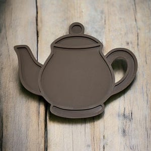May include: A brown teapot-shaped coaster with a dark brown outline. The coaster features a handle, spout, and a small knob on the lid. The coaster is made of a solid material and is placed on a wooden surface.