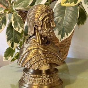 May include: A metallic gold-coloured bust sculpture depicting a person in profile with a textured beard, wearing an ornate headdress and a detailed garment with intricate patterns. The figure is mounted on a circular base with a decorative border.