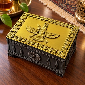Puede incluir: Una caja rectangular con tapa de metal dorado que presenta un emblema Faravahar en relieve y un borde de hojas. Los lados de la caja son de color gris oscuro con acabado mate, decorados con motivos florales y detalles en forma de gota.