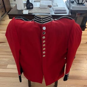 May include: A bright red military-style tunic featuring a high collar with black and white horizontal stripes, long sleeves with black cuffs, and a vertical row of silver-toned buttons running down the center front of the garment.