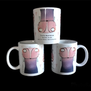 May include: Three white ceramic mugs with a cartoon illustration of a pink cat wearing a purple dress. The text on the mug reads "Tante Bartsch Katze Olga hat im März geheiratet."