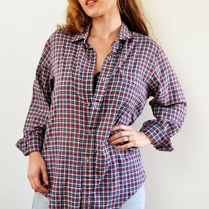 May include: A woman wearing a red and blue plaid flannel shirt with a black leather cap. She is wearing denim shorts and has her hand on her hip.