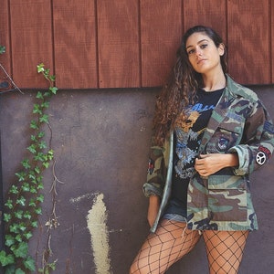 May include: A woman wearing a camouflage jacket with patches, a black t-shirt with a graphic print, fishnet stockings, and black boots. She is standing in front of a wooden wall and a gray wall with ivy growing on it.