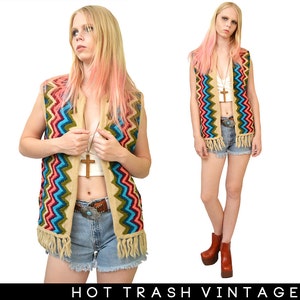 May include: A colorful crocheted vest with fringe, featuring a chevron pattern in shades of blue, green, pink, and red. The vest is worn over a white crop top and blue denim shorts.