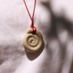 Könnte beinhalten: Eine cremefarbene Keramik-Anhänger-Halskette mit einem Spiraldesign. Der Anhänger hängt an einer dünnen, leuchtend roten Kordel. Die Halskette wird vor einem weißen Hintergrund gezeigt.