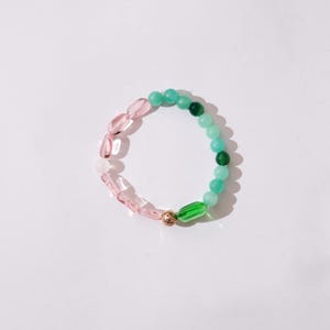 Könnte beinhalten: Ein Armband mit einer Mischung aus rosa und grünen Perlen. Das Armband hat ovale rosa Perlen, die in runde grüne Perlen übergehen. Eine goldfarbene Perle mit grünem Akzent ist ebenfalls vorhanden.